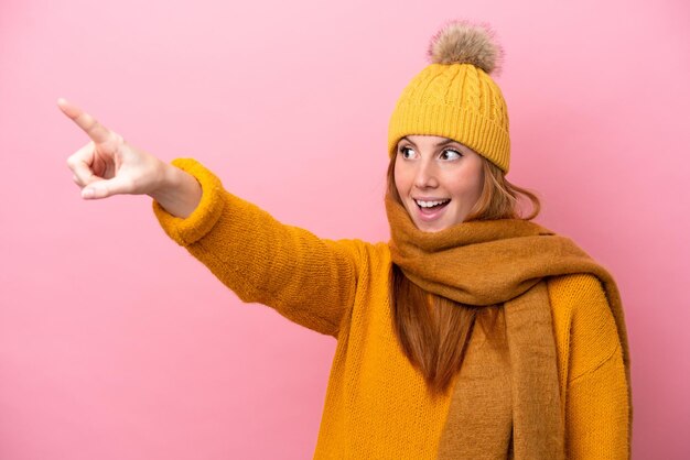 Jonge roodharige vrouw die een winterjas draagt die op een roze achtergrond wordt geïsoleerd en wegwijst