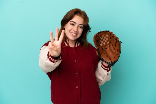 Jonge roodharige vrouw die honkbal speelt geïsoleerd op blauwe achtergrond gelukkig en telt drie met vingers