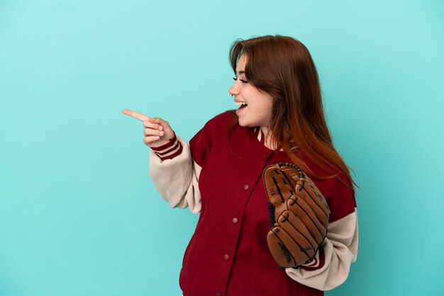 Jonge roodharige vrouw die honkbal speelt geïsoleerd op een blauwe achtergrond die met de vinger naar de zijkant wijst en een product presenteert