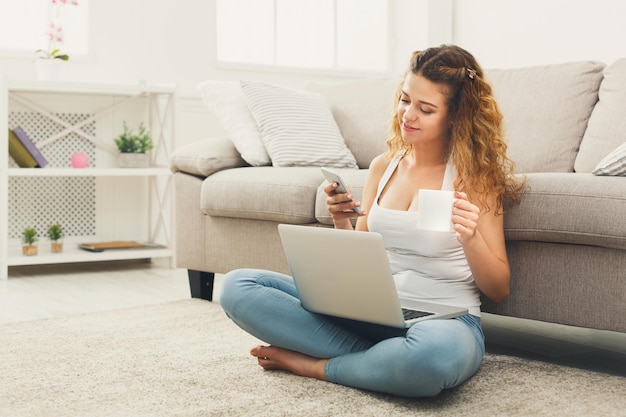 Jonge roodharige vrouw die smartphone controleert terwijl ze aan een laptop werkt, thuis op de vloer zit, kopieer ruimte