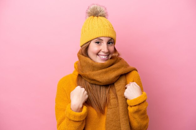 Jonge roodharige vrouw die winterjas draagt die op roze achtergrond met verrassingsgezichtsuitdrukking wordt geïsoleerd