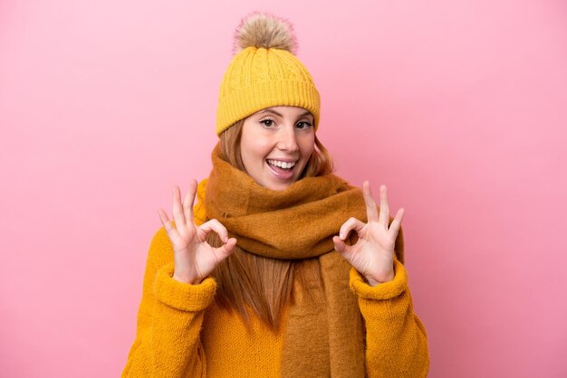 Jonge roodharige vrouw die winterjas draagt die op roze achtergrond wordt geïsoleerd en een ok teken met vingers toont