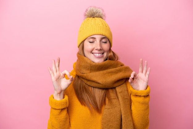 Jonge roodharige vrouw draagt winterjas geïsoleerd op roze achtergrond in zen pose