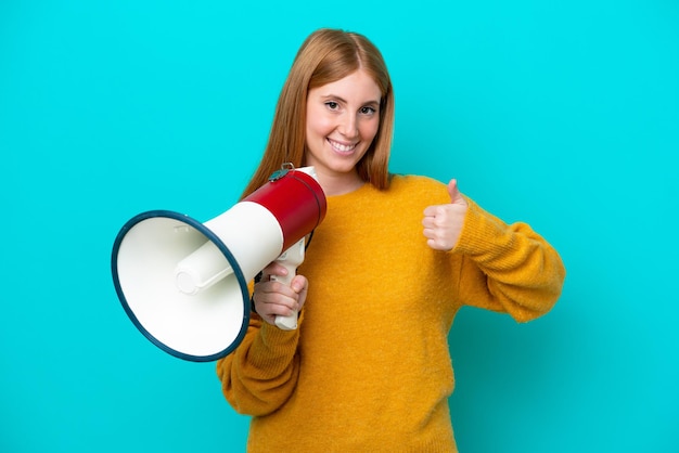 Jonge roodharige vrouw geïsoleerd op blauwe achtergrond met een megafoon met duim omhoog