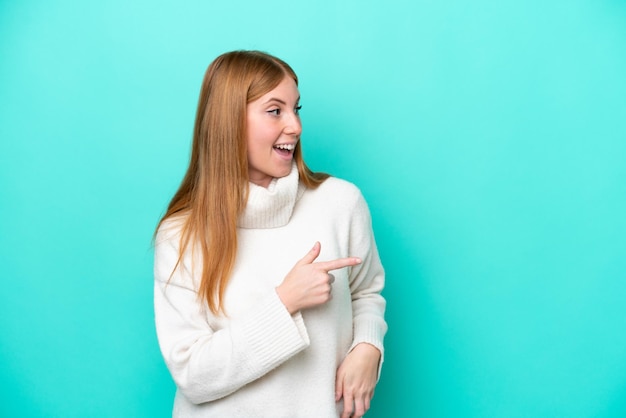 Jonge roodharige vrouw geïsoleerd op een blauwe achtergrond die met de vinger naar de zijkant wijst en een product presenteert