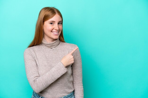 Jonge roodharige vrouw geïsoleerd op een blauwe achtergrond die naar de zijkant wijst om een product te presenteren