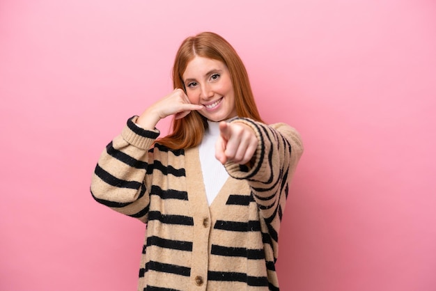 Jonge roodharige vrouw geïsoleerd op een roze achtergrond die een telefoongebaar maakt en naar voren wijst