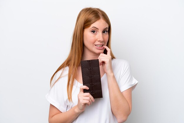 Jonge roodharige vrouw geïsoleerd op een witte achtergrond die een chocoladetablet neemt en twijfels heeft