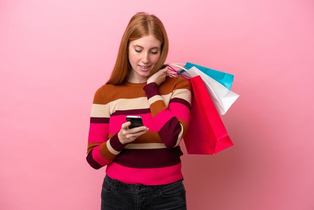 Jonge roodharige vrouw geïsoleerd op roze achtergrond met boodschappentassen en het schrijven van een bericht met haar mobiele telefoon naar een vriend