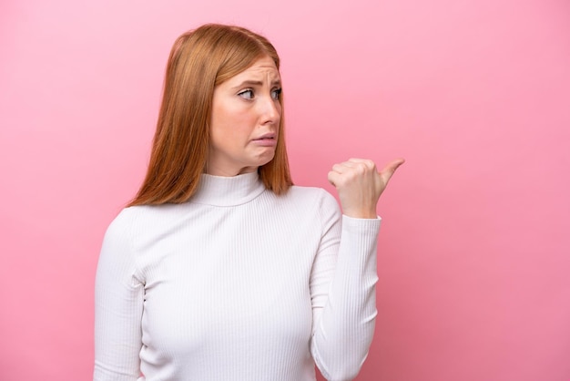 Jonge roodharige vrouw geïsoleerd op roze achtergrond ongelukkig en wijzend naar de zijkant