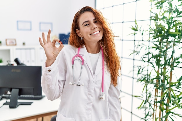 Jonge roodharige vrouw in doktersuniform en stethoscoop in de kliniek glimlachend positief doend ok teken met hand en vingers. succesvolle uitdrukking.