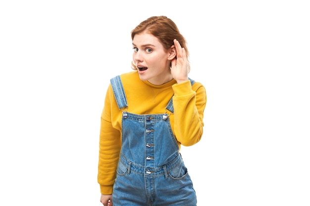 Jonge roodharige vrouw in vrijetijdskleding glimlachend en hand in de buurt van oor luisterend aandachtig horend geïsoleerd op witte achtergrond