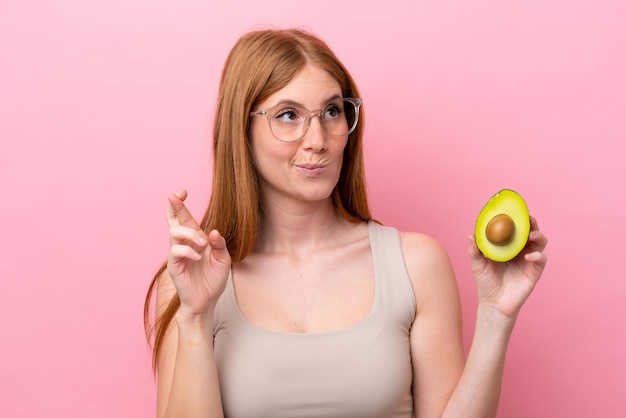 Jonge roodharige vrouw met een avocado geïsoleerd op een roze achtergrond met vingers die elkaar kruisen en het beste wensen