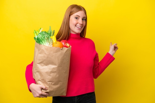Jonge roodharige vrouw met een boodschappentas geïsoleerd op een gele achtergrond die naar achteren wijst