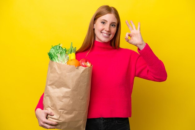 Jonge roodharige vrouw met een boodschappentas geïsoleerd op een gele achtergrond met ok teken met vingers