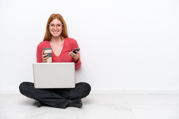 Jonge roodharige vrouw met een laptop zittend op de vloer geïsoleerd op een witte achtergrond met koffie om mee te nemen en een mobiel