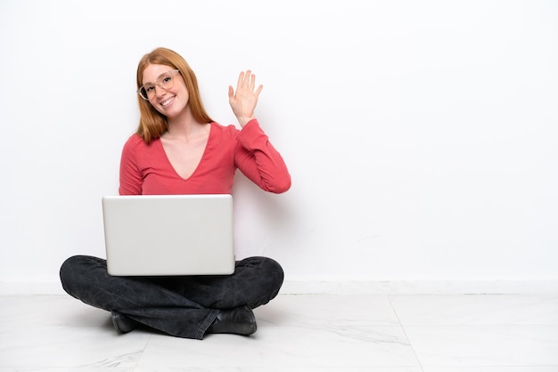 Jonge roodharige vrouw met een laptop zittend op de vloer geïsoleerd op een witte achtergrond saluerend met de hand met gelukkige uitdrukking