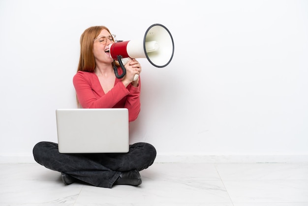 Jonge roodharige vrouw met een laptop zittend op de vloer geïsoleerd op een witte achtergrond schreeuwend door een megafoon