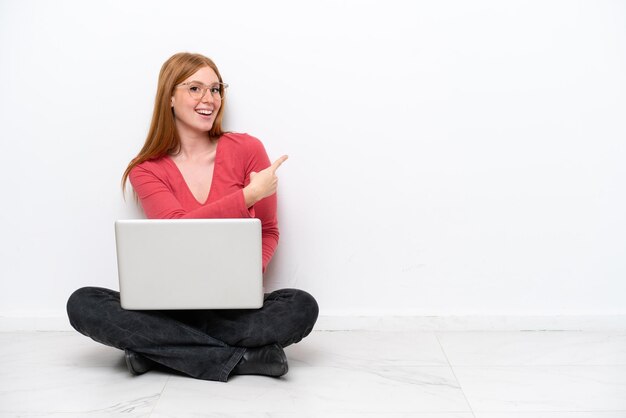 Jonge roodharige vrouw met een laptop zittend op de vloer geïsoleerd op een witte achtergrond wijzend naar achteren