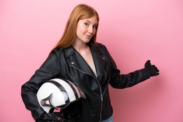 Jonge roodharige vrouw met een motorhelm geïsoleerd op een roze achtergrond die de handen naar de zijkant uitstrekt om uit te nodigen om te komen