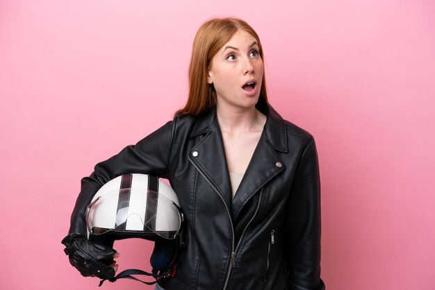 Jonge roodharige vrouw met een motorhelm geïsoleerd op roze achtergrond opzoeken en met verbaasde uitdrukking