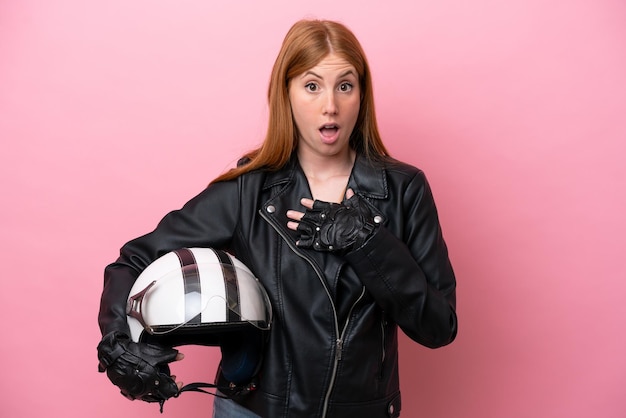 Jonge roodharige vrouw met een motorhelm geïsoleerd op roze achtergrond verrast en geschokt terwijl ze naar rechts kijkt