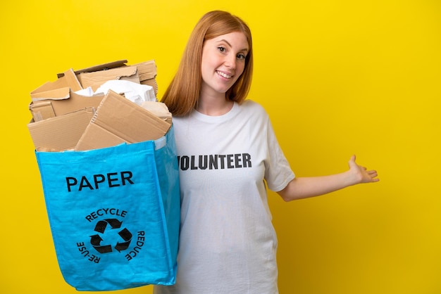 Jonge roodharige vrouw met een recyclingzak vol papier om te recyclen geïsoleerd op een gele achtergrond die de handen naar de zijkant uitstrekt om uit te nodigen om te komen