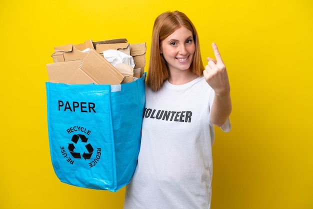 Jonge roodharige vrouw met een recyclingzak vol papier om te recyclen geïsoleerd op een gele achtergrond die een komend gebaar doet