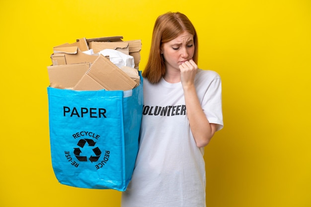 Jonge roodharige vrouw met een recyclingzak vol papier om te recyclen geïsoleerd op gele achtergrond met twijfels