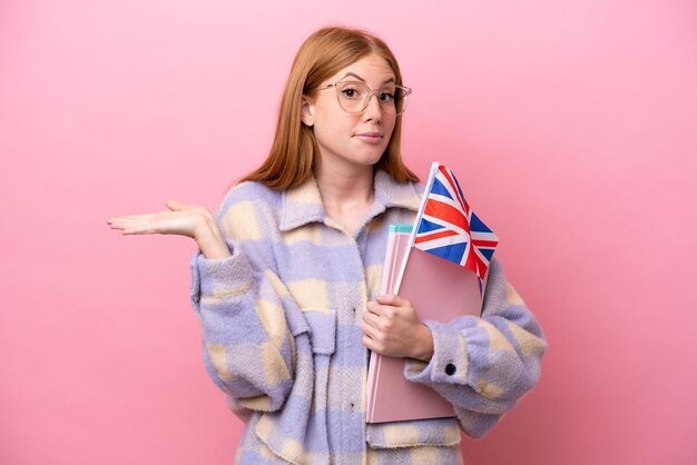 Jonge roodharige vrouw met een vlag van het verenigd koninkrijk geïsoleerd op een roze achtergrond die twijfelt terwijl ze haar handen opsteekt