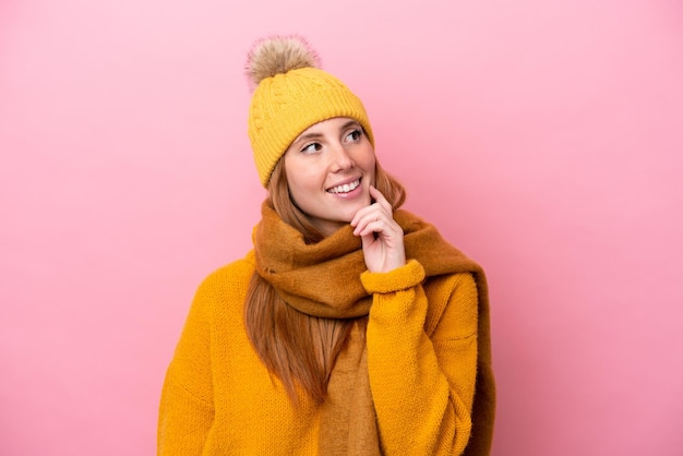 Jonge roodharige vrouw met een winterjas geïsoleerd op een roze achtergrond die een idee denkt terwijl ze omhoog kijkt