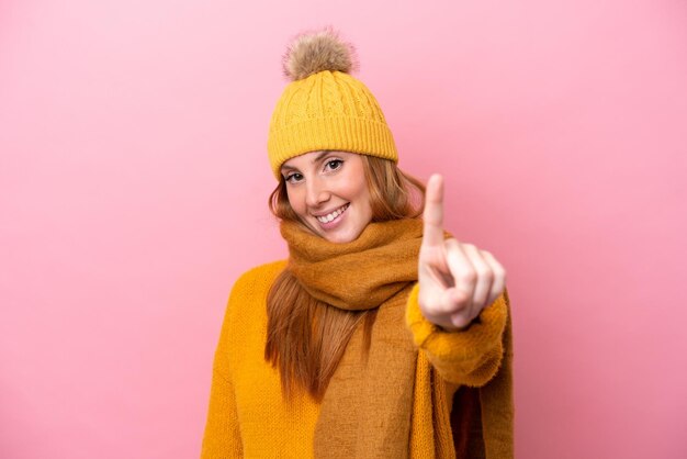 Jonge roodharige vrouw met een winterjas geïsoleerd op een roze achtergrond die een vinger laat zien en optilt