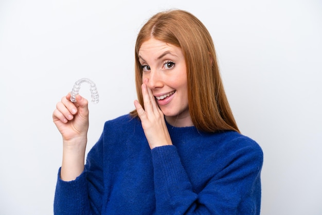 Jonge roodharige vrouw met onzichtbare beugels geïsoleerd op een witte achtergrond die iets fluistert