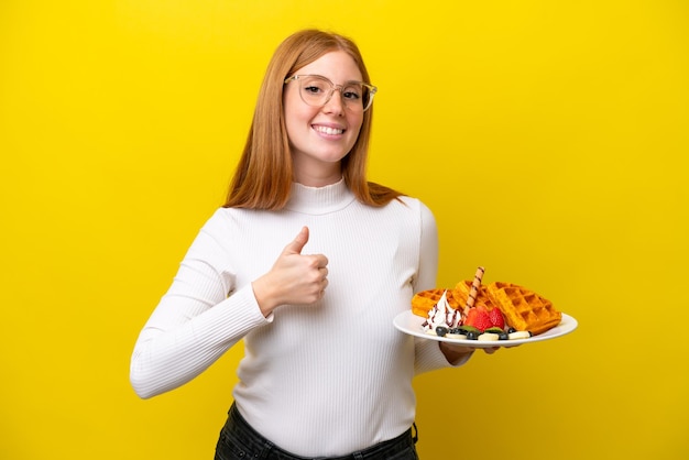 Jonge roodharige vrouw met wafels geïsoleerd op een gele achtergrond met een duim omhoog gebaar