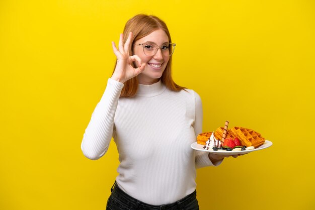 Jonge roodharige vrouw met wafels geïsoleerd op een gele achtergrond met ok teken met vingers