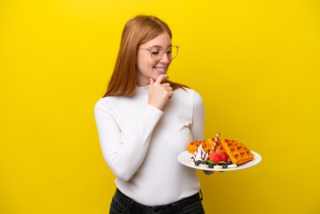 Jonge roodharige vrouw met wafels geïsoleerd op een gele achtergrond, opzij kijkend en glimlachend
