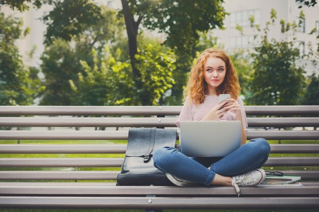 Jonge roodharige vrouw multitasking. Zittend op de bank in het park surfen op het web en chatten op mobiel. Freelancer werken zakenvrouw levensstijl concept