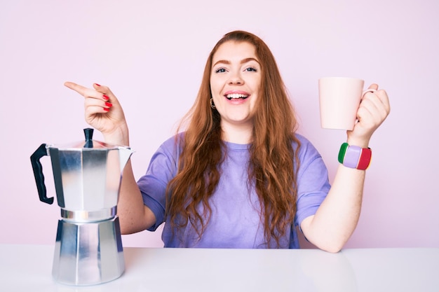 Jonge roodharige vrouw zit op de tafel en drinkt koffie met een gelukkige glimlach en wijst met haar hand en vinger naar de zijkant