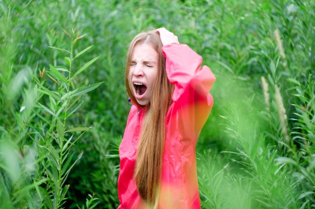 Jonge roodharigevrouw die in roze regenjas wegens saai bewolkt weer geeuwen