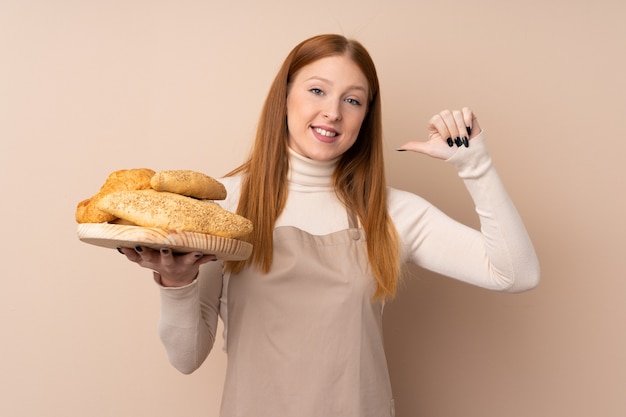 Jonge roodharigevrouw in eenvormige chef-kok. Vrouwelijke bakker die een tafel met verschillende broden trots en zelfvoldaan houdt