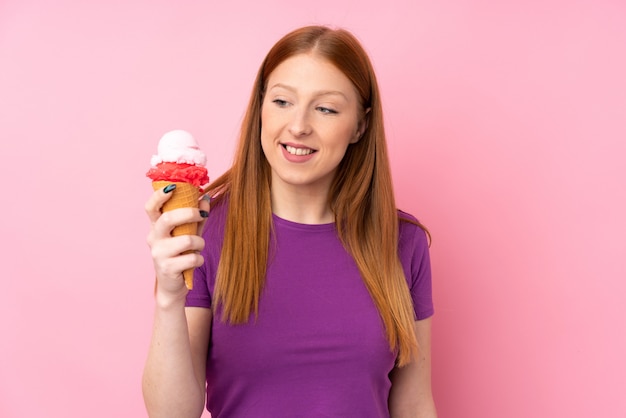 Jonge roodharigevrouw met een cornetroomijs over geïsoleerde roze muur met gelukkige uitdrukking