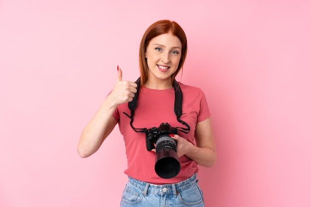 Jonge roodharigevrouw over geïsoleerde roze achtergrond met een professionele camera en met omhoog duim