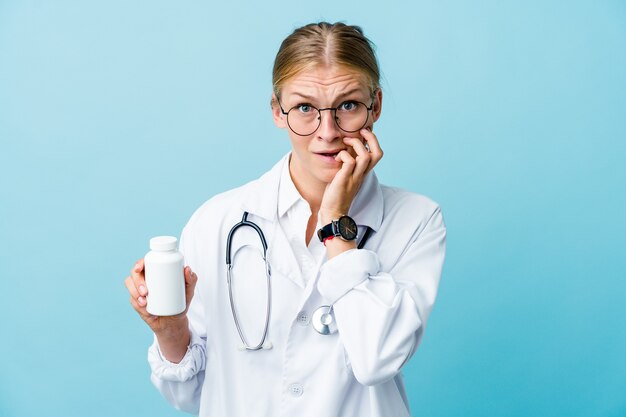 Jonge russische dokter vrouw met pillen fles op blauwe nagels bijten, nerveus en erg angstig.