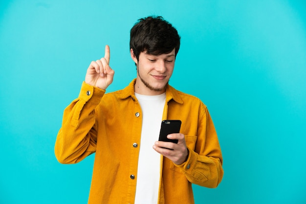 Jonge Russische man geïsoleerd op blauwe achtergrond met behulp van mobiele telefoon en het opheffen van de vinger