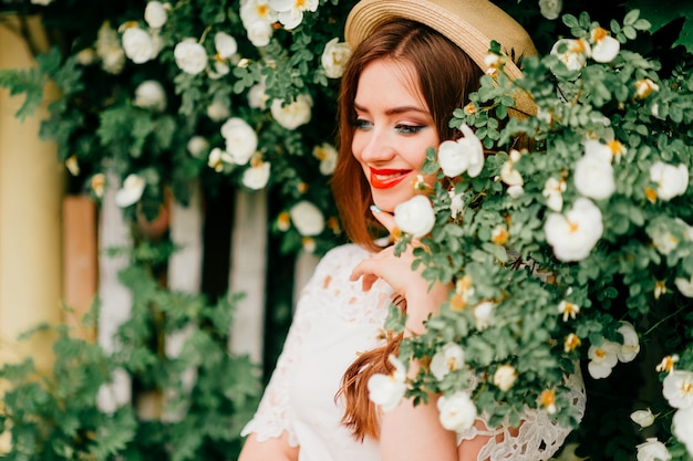 Jonge Russische schoonheid. Aantrekkelijk meisje in vintage top en krullend rood haar en strooien hoed poseren voor camera met hek en groene bomen