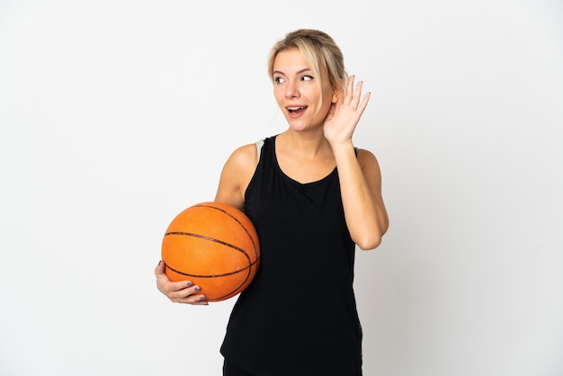 Jonge Russische vrouw die basketbal speelt geïsoleerd op een witte achtergrond die naar iets luistert door de hand op het oor te leggen