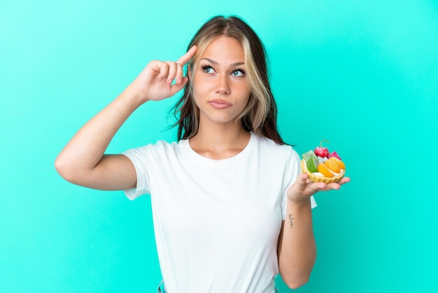 Jonge russische vrouw die een fruitzoet houdt die op blauwe achtergrond wordt geïsoleerd die twijfels heeft en denkt