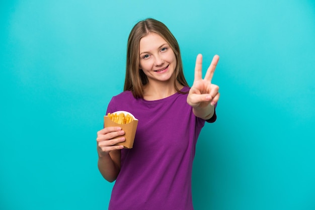 Jonge Russische vrouw die frietjes vangt die op blauwe achtergrond worden geïsoleerd en glimlacht en overwinningsteken toont