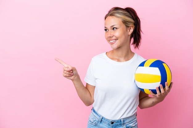 Jonge Russische vrouw die volleybal speelt geïsoleerd op een roze achtergrond die met de vinger naar de zijkant wijst en een product presenteert