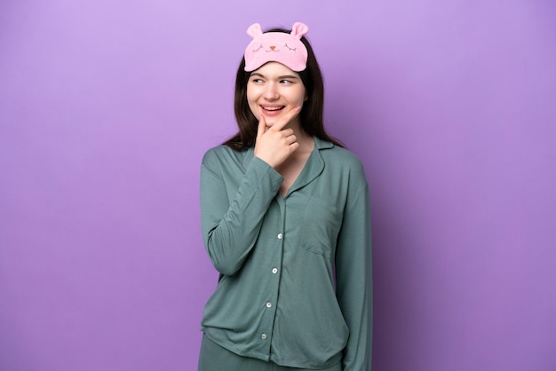 Jonge Russische vrouw in pyjama geïsoleerd op paarse achtergrond op zoek naar de kant en glimlachend